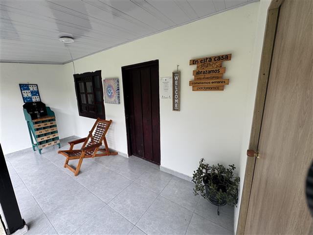 eco-hotel-gaia-21 - Hoteles en el Quindío Eje Cafetero, Alojamientos, Alquiler de fincas Covered porch area with a wooden lounge chair, potted plant, and a set of wall plaques including a Spanish sign saying 'En esta casa abrazamos cometemos errores aprendemos' near a dark doorway.