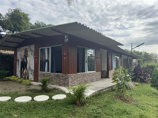 eco-hotel-gaia-3 - Hoteles en el Quindío Eje Cafetero, Alojamientos, Alquiler de fincas Single-story building with a brick base, wood siding, and a wide covered porch; large windows, a stepping-stone path, and tropical landscaping in front.