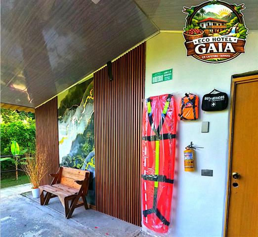 eco-hotel-gaia-6 - Hoteles en el Quindío Eje Cafetero, Alojamientos, Alquiler de fincas Outdoor hotel entrance area at Eco Hotel Gaia, with a wooden bench, vertical slats, and the Gaia logo on the wall; red life-vest flotation bags and a safety extinguisher mounted near a door.